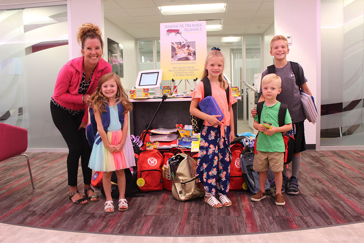 1ST SUMMIT BANK collects school supplies for kids showing 4 kids and bank employee surrounded by supplies