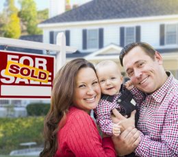 a man and woman holding a baby in front of a for sale sign.