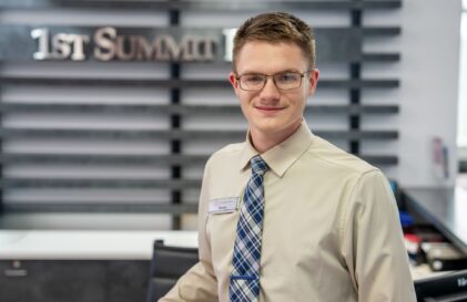 Young man in a beige shirt and plaid tie smiles at the camera in a modern reception area; name badge reads Devin in the foreground.
