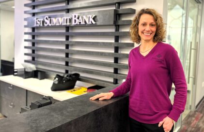 Woman in a magenta sweater stands at a bank reception desk, smiling, with a modern lattice wall and '1st Summit Bank' sign behind her